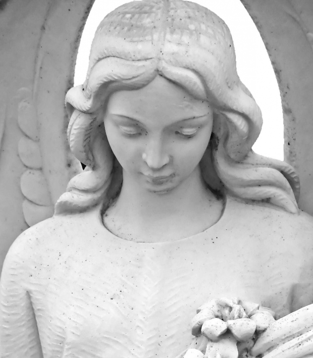 White statue of an angel, a woman with long flowing hair looking down and holding flowers in her arms