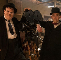 One large and one small man both in black suits with white shirts and hats leaning on an old time film projector