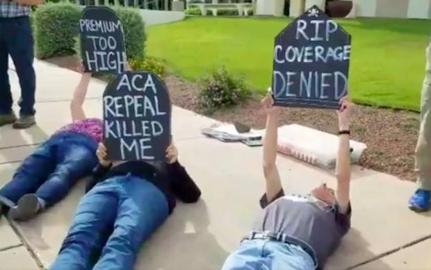 People laying outside on the ground holding tombstone signs that say ACA Repeal killed me
