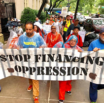 People wearing colorful clothes and raingear marching outside holding a banner reading Stop Financing Oppression