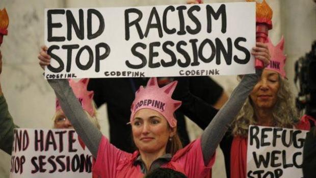 Blonde woman in pink with Code Pink crown holding a sign that says End racism Stop Sessions