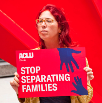 Woman with glasses and long red hair outside holding a sign that says Stop Separating Families