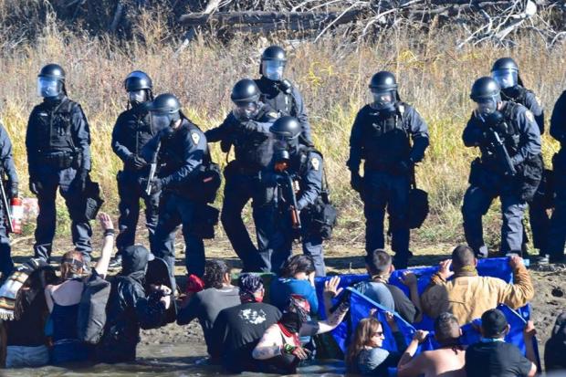Photo of heavily armed soldier-looking men pointing guns at Native American protestors sitting on the ground