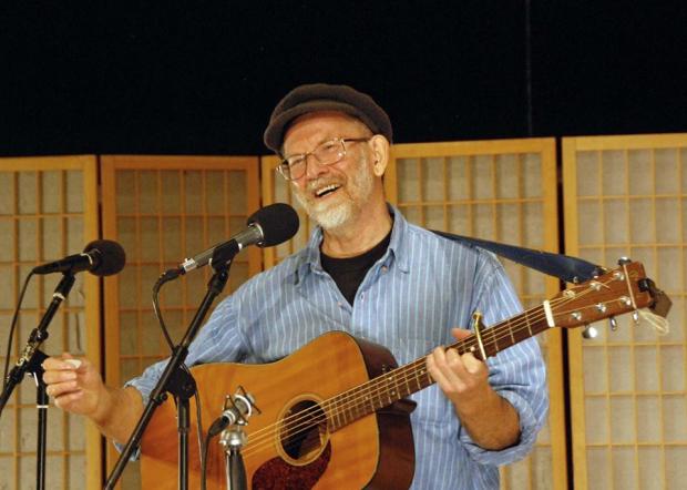 Man with gray goatee and black cap holding guitar at microphone smiling 