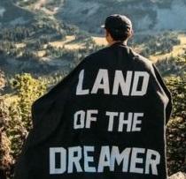 Man standing on a hill looking out with his back to the camera with a cape on with the words Land of the Dreamer