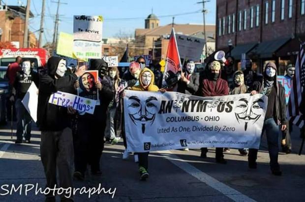 People marching wearing white and black Guy Fawkes masks carrying a long banner