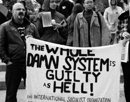 Black and white photo with people in winter coats outside gathered in a group behind a white banner with black letters saying The Whole Damn System is Guilty as Hell