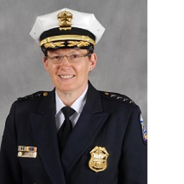 White woman smiling with wire-rimmed glasses wearing a large white police hat and a blue coat with a gold badge