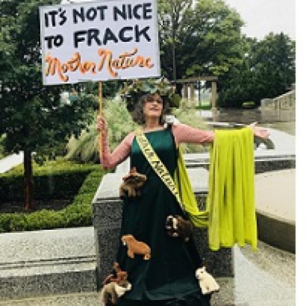 Woman in a long green dress with a sash and a cape and crown of flowers with a sign saying It's not nice to frack mother nature