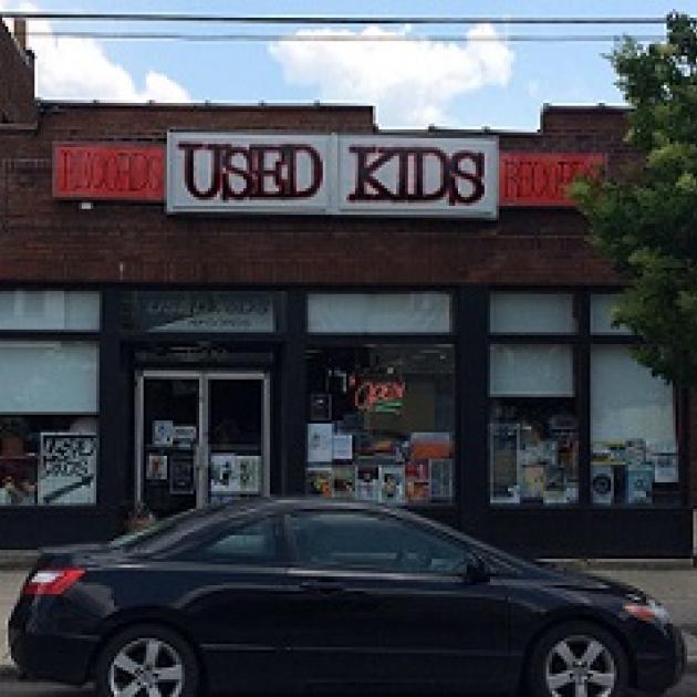 Brick building with sign at top saying Used Kids and a storefront with windows, a black car parked in front