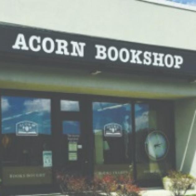 Front of a building with ACORN BOOKSHOP on the sign, windows and a front door of glass, flowers out front.