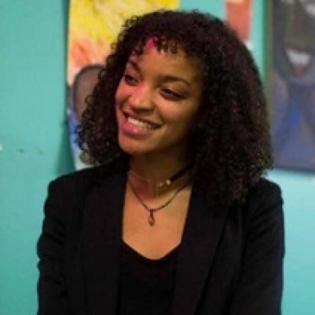 Young black woman smiling turning her head to the left wearing a black suit against a colorful wall of art