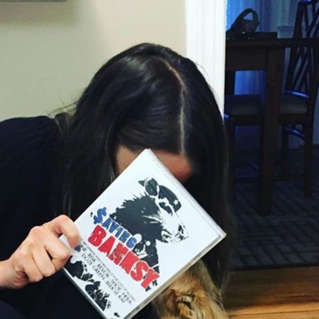 Women with dark hair leaning over hiding her face with a book, Saving Banksy