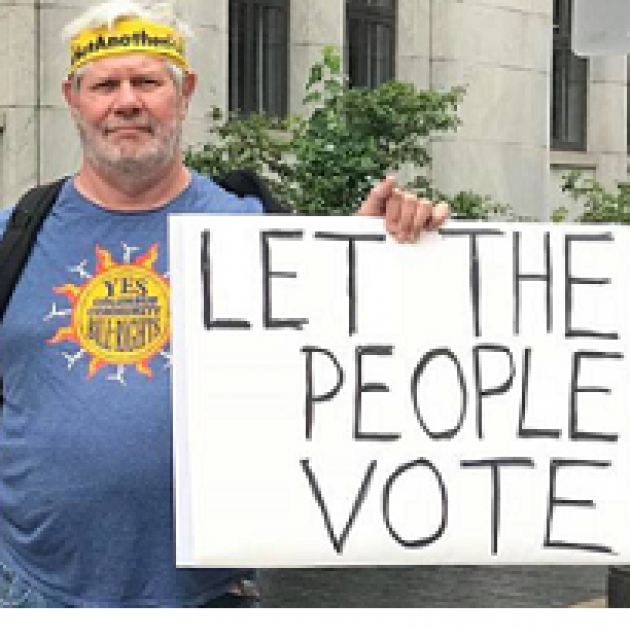 White man with yellow headband and blue T-Shirt with sun in the middle holding a sign that says Let the People Vote