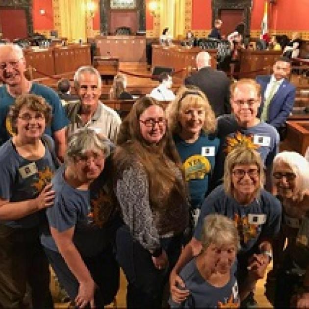 Lots of smiling white people male and female leaning toward each other all wearing blue T-shirts in a city council chambers