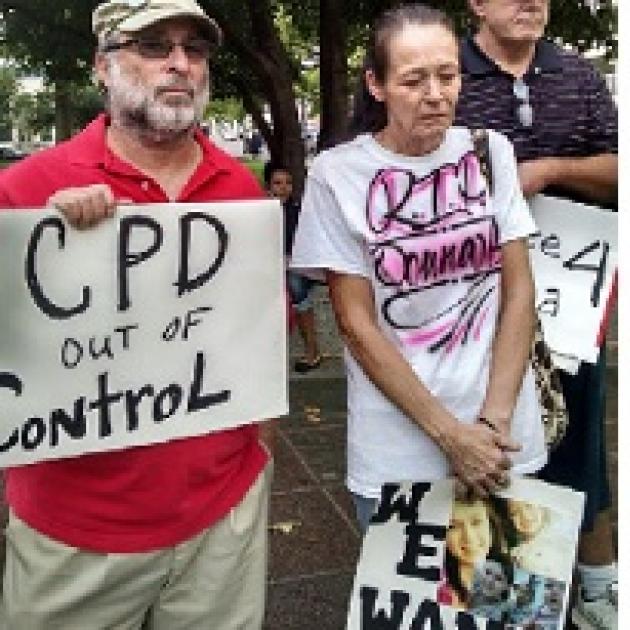 Man with gray beard and sunglasses wearing a baseball cap holding a sign that says CPD out of control standing next to an older woman with gray hair pulled back in a ponytail looking down at the ground, very sad, wearing a white T-shirt with words "RIP Donna" on it and holding a sign with pictures of a young woman on it