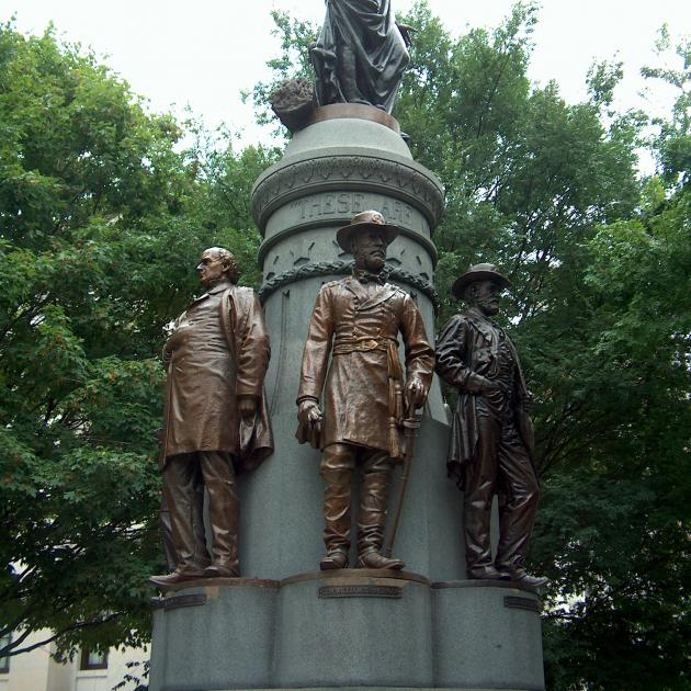 Gray pointy statue with Union Soldier figures standing with their backs around it against trees in the background