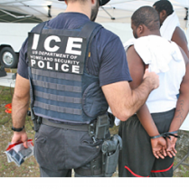Large white man in uniform that says ICE Police on the back with his back to the camera holding the arm of a dark skinned man in a white t-shirt and shorts with his back to the camera and his hands behind him in handcuffs