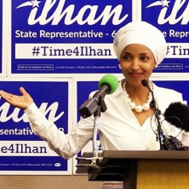 Young woman of color wearing a white hat and suit and pearl necklace standing in front of mics at a podium and signs with her name Ilhan State Representative in the background