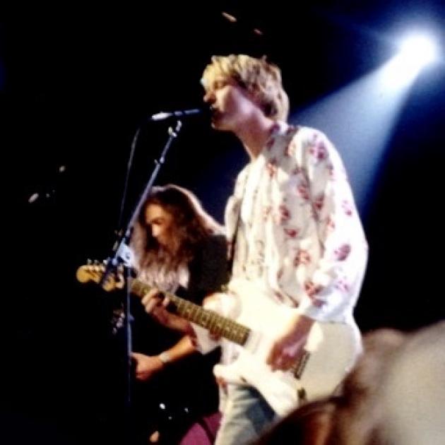 Kurt Cobain singing into a mic and a guitarist behind him