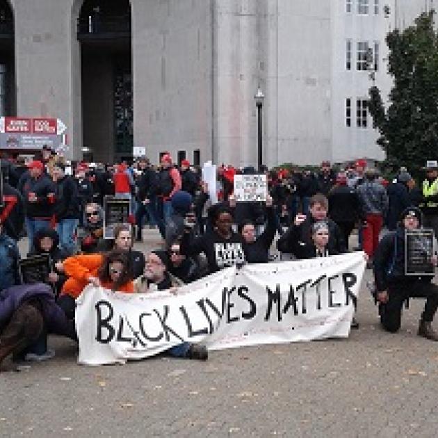 People wearing winter clothes bending down on one knee outside holding a long white banner with black letters reading Black Lives Matter in front of a large white building