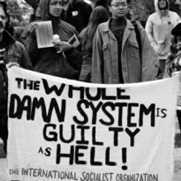 Black and white photo with people in winter coats outside gathered in a group behind a white banner with black letters saying The Whole Damn System is Guilty as Hell