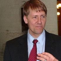 White man in a dark suit, white shirt, red tie with shaggy blonde hair with a pensive worried look
