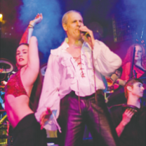 Middle aged white man with balding head wearing a blousy tie-front white shirt with tight shiny dark pants with a woman dancing sideways wearing a red bikini top and low-rider black pants and others dancing behind