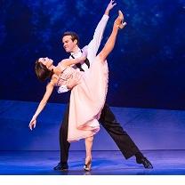 White man with dark hair in white shirt and black pants dancing with a white woman with short dark hair in a pick dress, he is leaning over as he holds her as she is kicking her leg high up in the air 