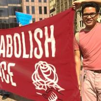 Young dark haired man with glasses holding a large red sign that says Abolish ICE with a DSA symbol of fist and rose