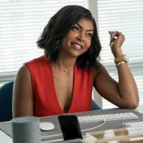 Young black woman smiling and sitting at a desk with computer with one elbow on the desk and arm up by her face in front of a window