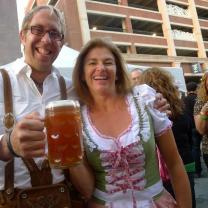 White ma with gray hair and glasses smiling holding a huge mug of beer with a woman who is wearing a German-looking corset and frilly top