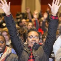 Black woman with glasses in front of a huge crowd of people smiling with both hands held high above her head