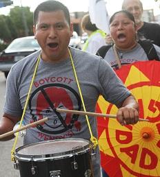 Latino man in foreground with drum around his neck and hitting it with drumsticks, his mouth open chanting, wearing a shirt that says Boycott Wendys with a picture of a girl's head with red pigtails and a No sign around it (circle with line through it) and a Latino woman behind him holding a bright orange and yellow sign also with her mouth open chanting