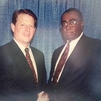 White middle aged man in a suit posing shaking hands with black man with glasses in a suit