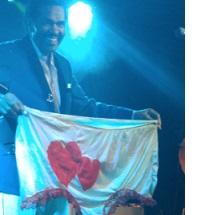 Smiling black man standing in a suit on a stage holding up a huge pair of women's undies that are white with two red harts on them