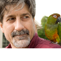 White man with grayish hair and beard with green parakeet on his shoulder