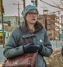 Young woman with brown rimmed glasses, long brown hair in winter clothes outside with her purse over her shoulder an holding it close to her body, looking like she is talking