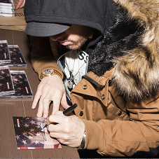 Guy in a black hoodie and brown heavy winter coat with fur collar sitting at a table with his face looking down where he is signing a CD