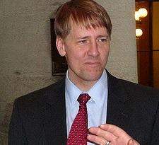 White man in a dark suit, white shirt, red tie with shaggy blonde hair with a pensive worried look
