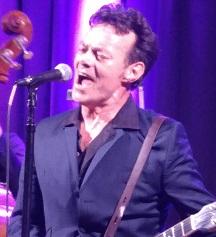White man at a microphone belting out a song wearing a dark suit and shirt unbuttoned with a guitar strapped around him
