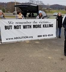 People outside holding a long white banner with black letters saying We remember the victims...But not with more killing and www.abolition.org and a phone number