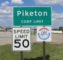 Green road sign saying Piketon Corp Limit, another sign saying Speed Limit 50 and a sign with an Ohio logo on it and in the background a highway and some grass and the blue sky with white clouds