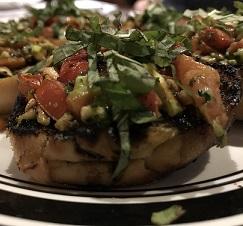 A piece of food on a plate with other similar items, looks like a baked potato with lots of greens, tomatoes, other brownish things on top.