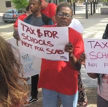 Black woman outside at rally holding a sign that says Tax $ for Schools Not for Scabs
