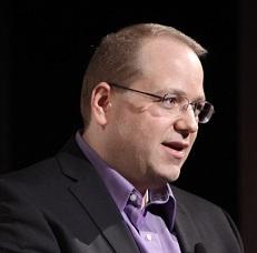 Middle aged white man with brown receding hair with wire rimmed glasses looking to the right with  a gray suitcoat and purple shirt his mouth open as if talking