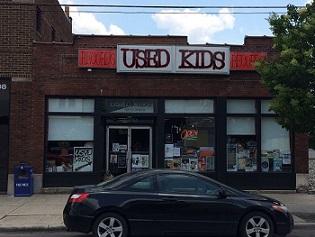 Brick building with sign at top saying Used Kids and a storefront with windows, a black car parked in front