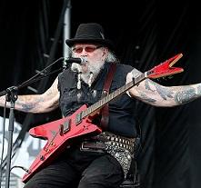 Older man with black hat, white hair and beard, sunglasses, sitting on a chair on stage with his arms spread wide with lots of tattoos and a red rock and roll electric guitar on his lap