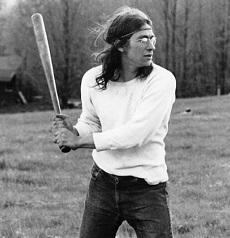 Black and white photo of a young man with long flowing brown hair with a headband across his forehead holding a baseball bat as if he's about to swing