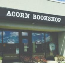 Front of a building with ACORN BOOKSHOP on the sign, windows and a front door of glass, flowers out front.